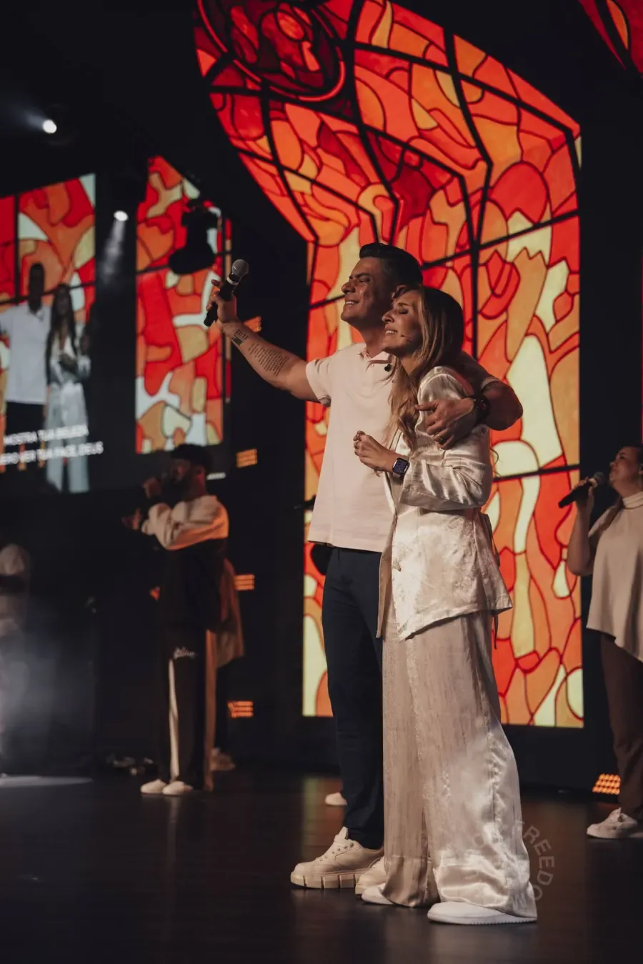 Pr. Cyllas e Pra. Thaís Marins abraçados no palco, diante do vitral vermelho